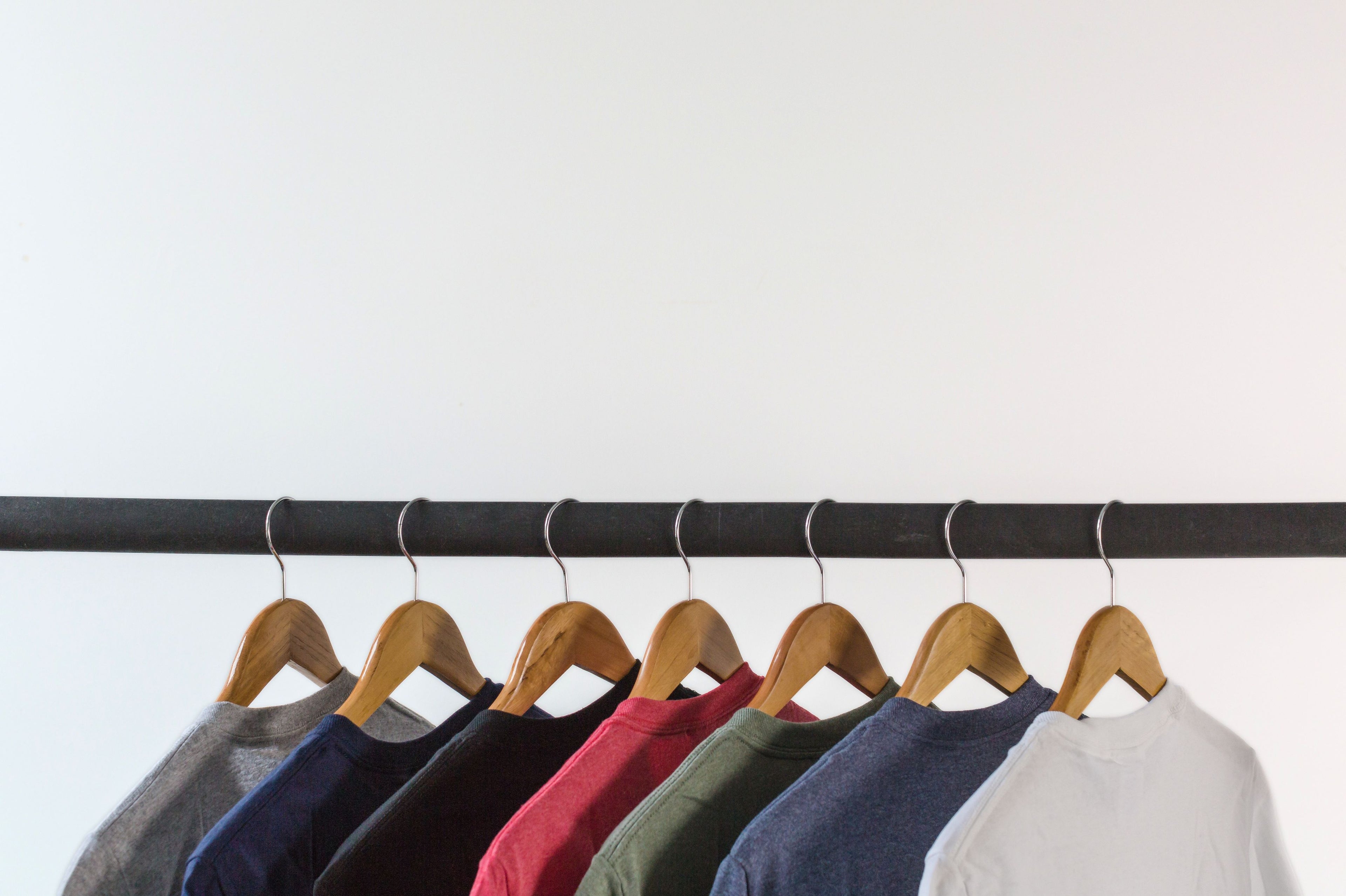 A row of crewneck sweatshirts in multiple colors hanging on wooden hangers from a black clothing rack against a plain white wall.